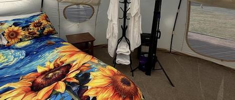 A tented room with a bed featuring a sunflower print, a small table, a clothing rack, and a window with a view.