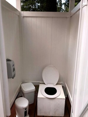 Uma pequena casa de banho exterior fechada, com fachada branca, assento sanitário e dispensador de álcool em gel.