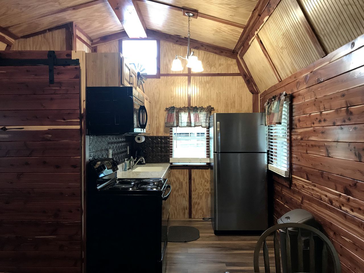 A compact kitchen with wooden walls, a stove, a refrigerator, and a window with blinds.