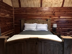 A wooden-framed bed with two pillows, a bedside table with a lamp, and a wooden ceiling.