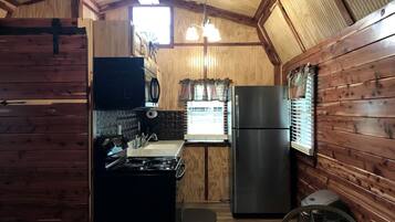 A compact kitchen with wooden walls, a stove, a refrigerator, and a window with blinds.