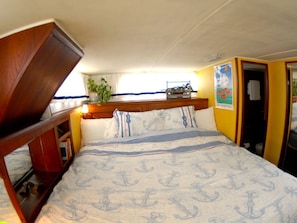 A compact room with a bed featuring a nautical anchor pattern, a small shelf with a plant, and a door leading to another room.