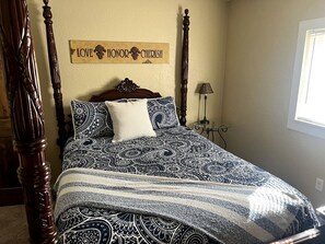 Un lit à baldaquin avec une couvre-lit et des coussins à motifs, une tête de lit en bois et un panneau mural portant les mots « AIMER, HONOURER, CHÉRIR ».