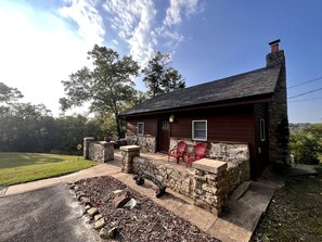 A stone building with a dark roof, a stone porch with red chairs, and a stone wall with a fire pit. - Cozy Cabin Rental near Table Rock Lake in the Ozark Mountains, Missouri (Hollister)