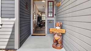 Una escultura de madera de un oso sosteniendo un cartel con la palabra 'Welcome'. En la entrada de un edificio, hay un felpudo y una puerta con marco rojo.