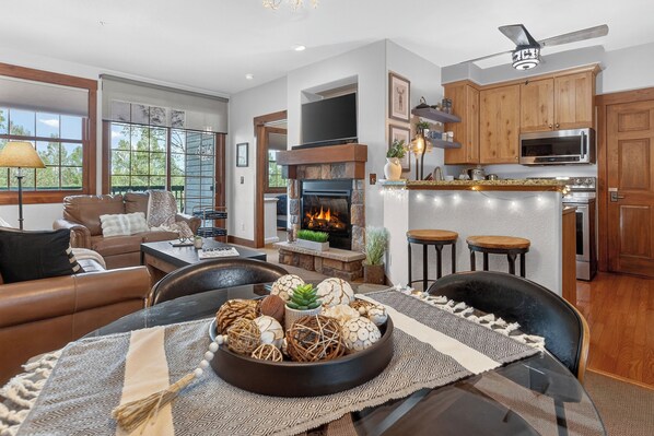 A modern living room with a fireplace, dining area, and kitchen with wooden cabinets and a flat-screen TV.