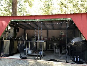 Een overdekte buitenkeuken met een rood dak, metalen tafels en lichtslingers.