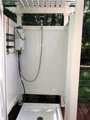 Une cabine de douche extérieure en bois peinte en blanc, équipée d’un tuyau d’arrosage et d’un boîtier de commande.