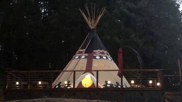 A tipi-style structure with a central light source, surrounded by a fence and trees.