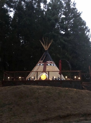 Tipi-tyyppinen rakennelma, jonka keskellä on valonlähde, ja jonka ympärillä on aita ja puita.