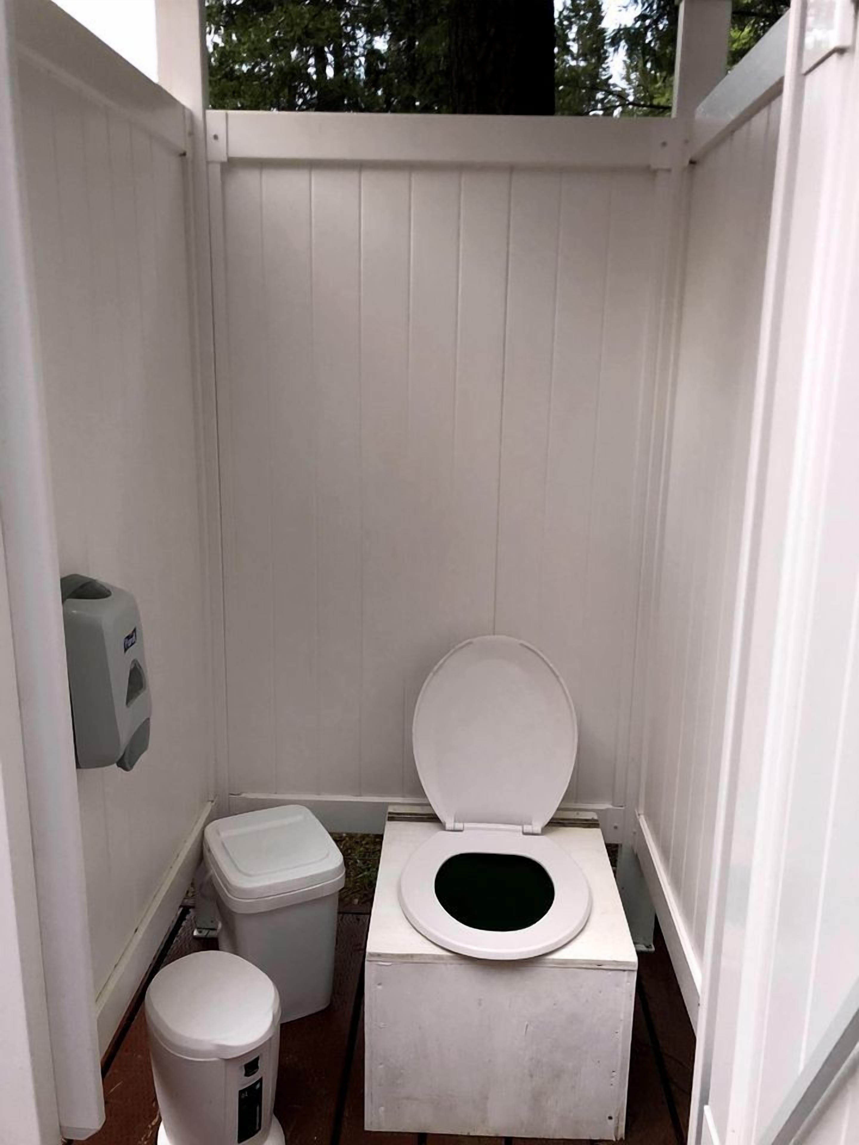A small, white, enclosed portable toilet with a lid, a waste bin, and a hand sanitizer dispenser.