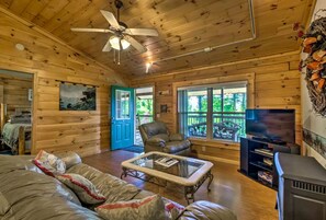 A wooden cabin interior with a ceiling fan, a sofa, a glass-top coffee table, a television, and a fireplace.
