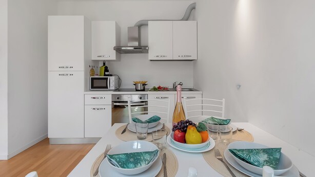 A modern kitchen with white cabinetry, a microwave, and a dining area with a table set for two. - Mediterranea Apartments (Savona)