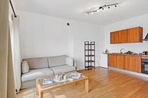Una sala de estar con un sofá, una mesa de centro con revistas, una cocina con alacenas de madera y un lavavajillas.