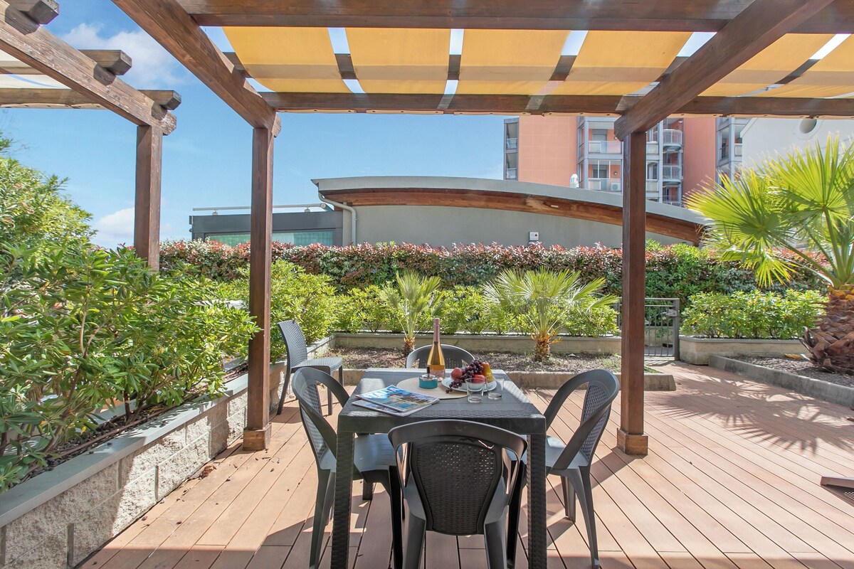 A covered outdoor patio with a table and chairs, surrounded by plants and a view of buildings.