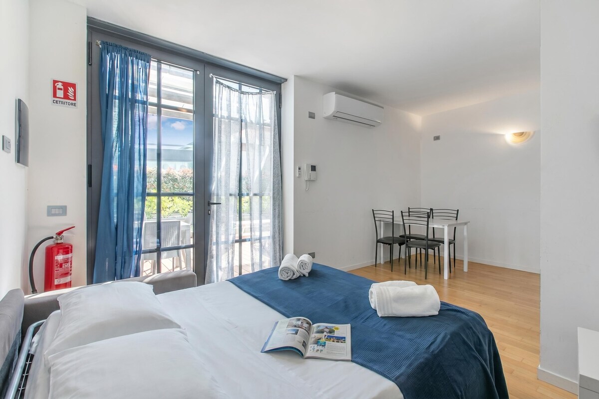 A hotel room with a bed, a table with chairs, a fire extinguisher, and a window with curtains.