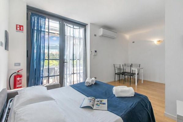 A hotel room with a bed, a table with chairs, a fire extinguisher, and a window with curtains. - Mediterranea Apartments (Savona)
