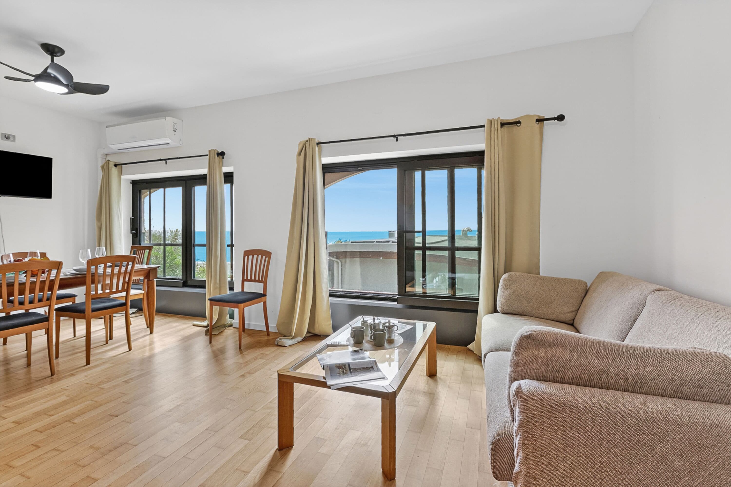 Ein großzügiger Wohnbereich mit Holzboden, einem Glastisch, einem beigen Sofa und einem Esstisch. Große Fenster bieten Blick auf das Meer.