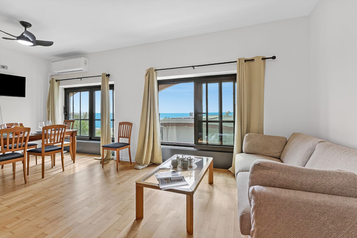 A spacious living area with a wooden floor, a glass-top coffee table, a beige sofa, and a dining set. Large windows offer a view of the sea.