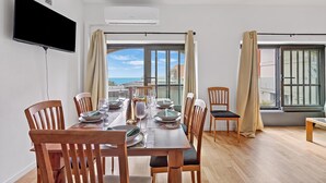 A dining area with a table set for four, wooden chairs, a flat-screen TV mounted on the wall, and a view of the sea through large windows. - Mediterranea Apartments (Savona)