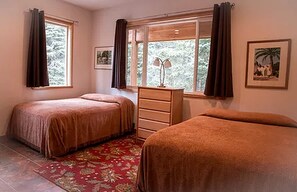 Ein Schlafzimmer mit zwei Betten, einem Schminktisch, einem Fenster mit Vorhängen und einem gerahmten Bild an der Wand.