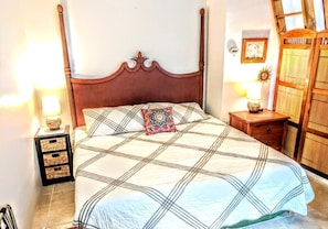 A bed with a wooden headboard, a patterned bedspread, and a decorative pillow. There are two bedside tables with lamps and a small drawer unit.