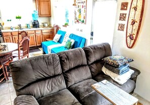 Ein Wohnzimmer mit einem dunkelbraunen Sofa, einem blauen Couchsofa und einem Couchtisch. Es gibt eine Küchenzeile mit Holzausbau und einem Esstisch.