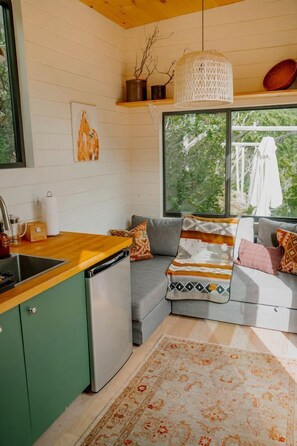 A compact kitchen with a dishwasher, a gray sofa with patterned cushions, and a large window offering a view of greenery.