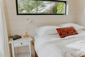 A bedroom with a bed, a nightstand with a lamp, a book, and a window with a view of trees.