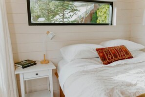 A bedroom with a bed, a nightstand with a lamp, a book, and a window with a view of trees.