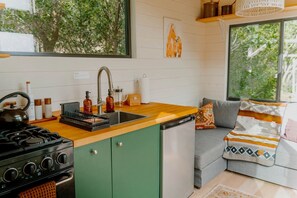 A compact kitchen with a sink, stove, and dishwasher, a green cabinet, and a window overlooking greenery.