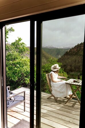 A person seated at a table with a bottle, overlooking a scenic mountainous landscape.