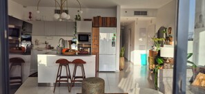 A modern kitchen with a central island, bar stools, a dining table, and a living area with a sofa and plants. - Bright & Stylish Boho Home w\ Botanical Terrace, by a grove -with a safe Mamad (Tel Aviv-Yafo)