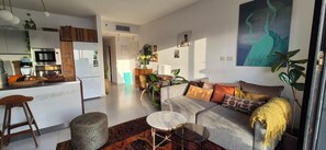 A modern living space with a kitchen, dining area, and a sofa with cushions. - Bright & Stylish Boho Home w\ Botanical Terrace, by a grove -with a safe Mamad (Tel Aviv-Yafo)