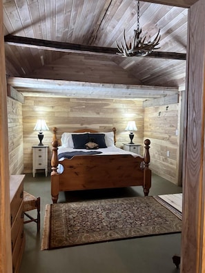 A wooden-paneled bedroom with a bed, two bedside tables with lamps, a rug, and a chandelier.