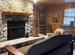 A cozy cabin interior with a stone fireplace, a woven basket, a chair, a ladder, and a window with a view of trees.