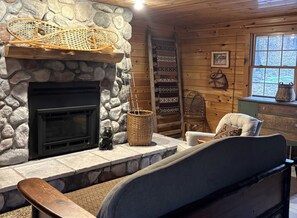 Ein gemütliches Blockhaus-Interieur mit Kamin aus Stein, einem gewebten Korb, einem Sessel, einer Leiter und einem Fenster mit Blick auf Bäume.