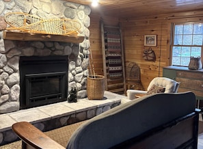Interior acogedor de una cabaña con chimenea de piedra, una cesta tejida, una silla, una escalera y una ventana con vistas a árboles.