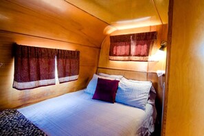 A small, cozy room with a bed, two windows with curtains, and a wall-mounted light. - Superb Elevated Trailer for Unique Vacation in San Bernardino County, California (Joshua Tree)
