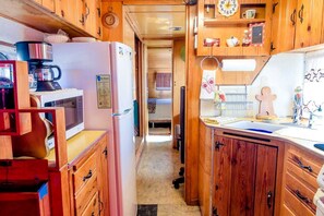 A narrow kitchen with wooden cabinets, a refrigerator, a coffee maker, and a clock on the wall. - Superb Elevated Trailer for Unique Vacation in San Bernardino County, California (Joshua Tree)