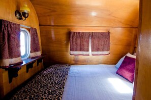 Une chambre confortable dans un camping-car, avec un lit, deux fenêtres équipées de rideaux, un banc et une lampe.
