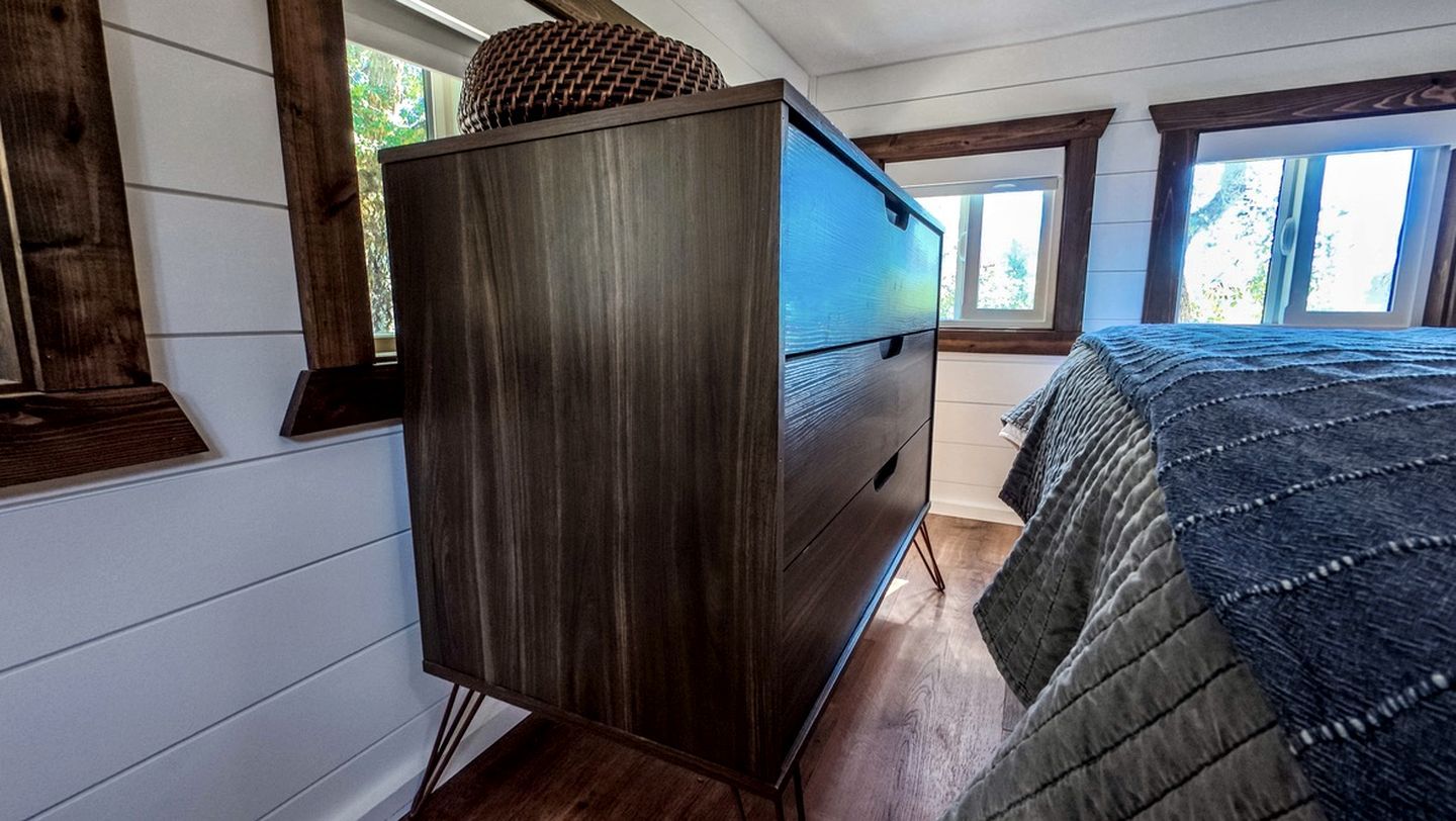 A bedroom with a dark wooden dresser, a bed with a quilt, and a window with a view of greenery.