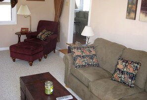 A living room with a red armchair, a beige sofa with patterned cushions, a small wooden table with a jar and remote control, and a lamp on a side table. - Charming Cottage Overlooking Gorgeous Citation Bass and Bream Pond near Charlottesville (Gordonsville)
