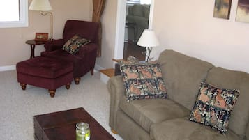 Ein Wohnzimmer mit einem roten Sessel, einem beigen Sofa mit gemusterten Kissen, einem kleinen Holztisch mit einer Glaseinlage und einer Fernbedienung sowie einer Lampe auf einem Beistelltisch.