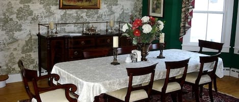 Un comedor con una mesa dispuesta para seis personas, rodeada de sillas de madera, un papel tapiz estampado, una pared de acento verde y una ventana con cortinas.