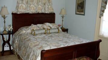A bedroom with a wooden bed, floral-patterned bedding, two bedside tables with lamps, a window with curtains, and a framed picture on the wall.