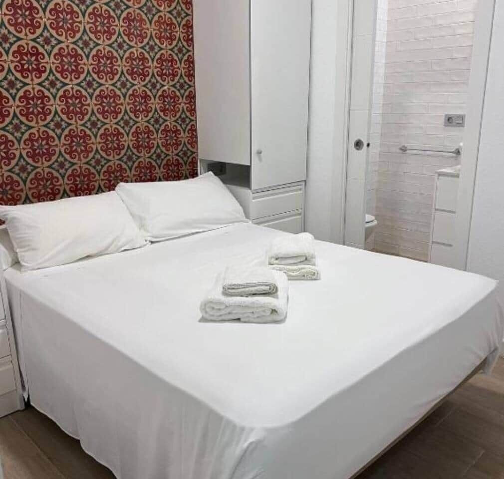 A neatly made bed with white linens and towels, a wardrobe, and a bathroom with a white tiled wall.