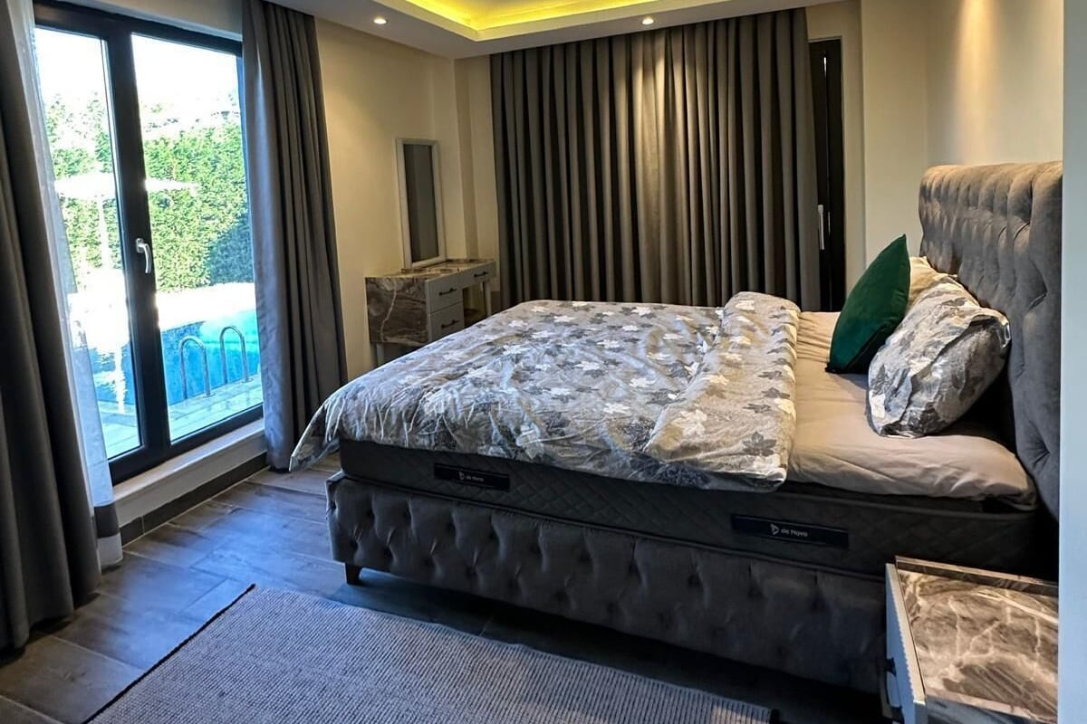 Une chambre moderne avec un grand lit, une petite table de chevet, une chaise, une vue sur l’extérieur et un plafond doté d’un éclairage encastré.