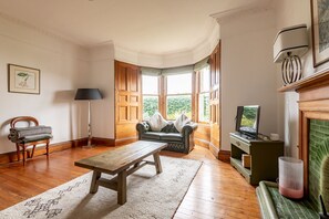 A room with a wooden floor, a sofa, a table, a lamp, a television, and a fireplace. - Woodburn Terrace (No 8) | St Andrews (St Andrews)