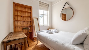 Un dormitorio con un escritorio de madera, una silla, una cama con ropa de cama blanca, un espejo redondo y una ventana con persianas.